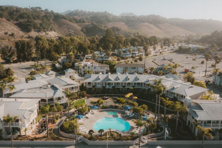Aerial view of a resort with a central pool, surrounded by palm trees and white buildings, set against hills and greenery under a hazy sky.