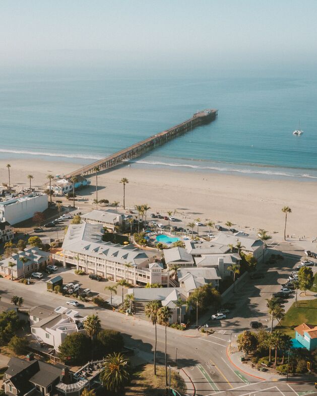 Aerial view of a beach town with a long wooden pier stretching into the ocean, sandy beach, palm trees, buildings, cars, and a pool, on a clear, sunny day.