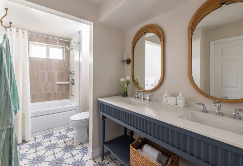 Bright bathroom with patterned floor tiles, double vanity with two sinks, wooden-framed mirrors, a basket with towels below, and a bathtub with a tiled shower and white curtain. White walls and natural light from a window.