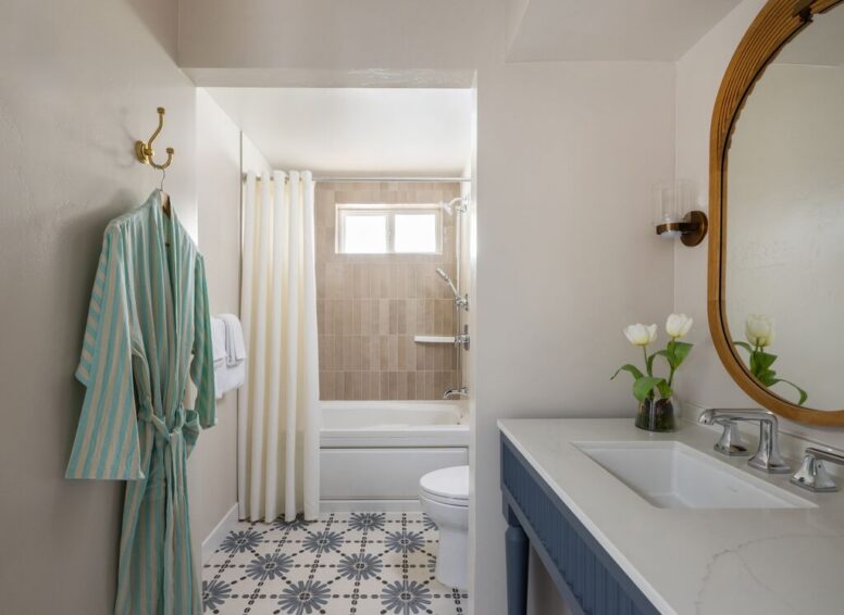 Bathroom with patterned tile floor, a white bathtub with beige wall tiles, a striped robe hanging on a hook, white sink with vase of tulips, and a round mirror above the sink.