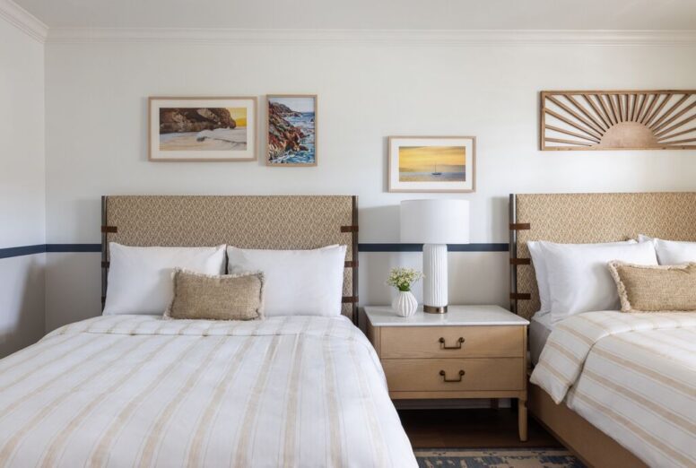 Two neatly made beds with beige-striped bedding flank a wooden nightstand with a white lamp, small vase, and books. Three landscape artworks hang on the white wall above the beds.
