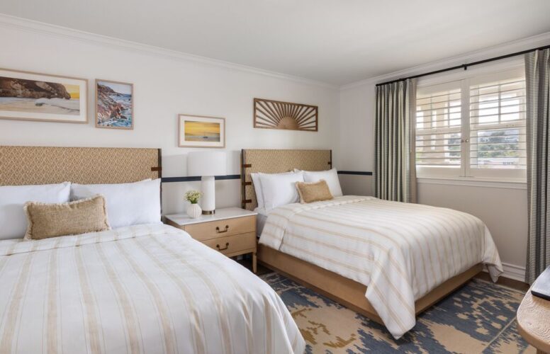 A bright bedroom with two beds, beige-striped bedding, a wooden nightstand, table lamp, artwork on the wall, and a window with white shutters and patterned curtains.