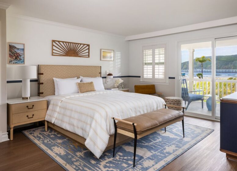 Bright bedroom with a king bed, striped bedding, two nightstands, a blue-patterned rug, a bench, armchair, and sliding doors leading to a balcony with a scenic waterfront view.
