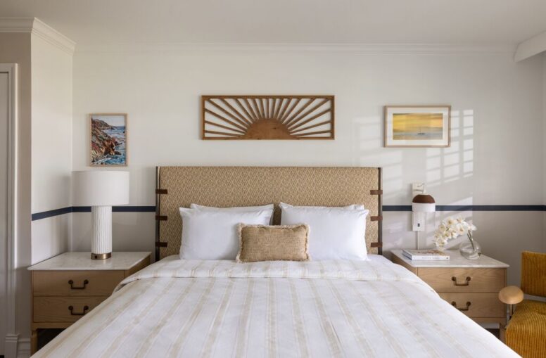 A neatly made bed with white linens, two pillows, and a tan headboard is centered between two nightstands. Art and lamps are on each nightstand, with framed artwork hung above the bed on a light-colored wall.