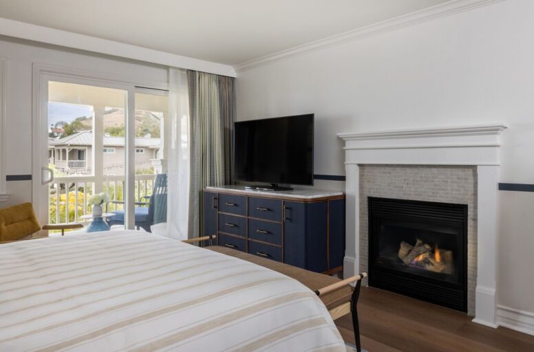 A cozy hotel room with a bed, striped bedding, a TV on a blue dresser, a lit fireplace, and a balcony with outdoor seating and a scenic view.
