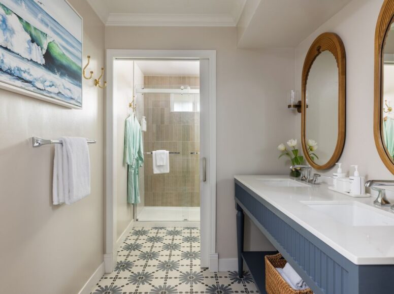 Bright bathroom with patterned tile floor, double sink vanity with two mirrors, towel and robe on hooks, shower with glass door, and a beach-themed painting on the wall.