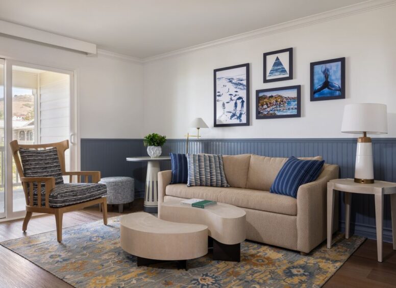 Modern living room with beige sofa, blue and beige pillows, wooden chair, round tables, abstract coffee table, blue-patterned rug, and framed wall art. Sliding glass door lets in natural light.