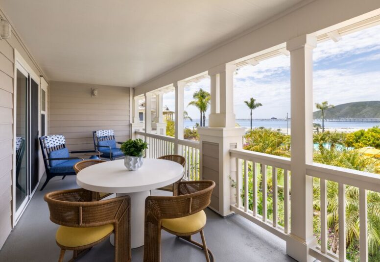 A covered balcony with a round white table, four wicker chairs, two lounge chairs, potted plant, and a view of palm trees, pool, and distant hills under a partly cloudy sky.