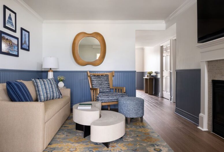 A cozy living room with a beige sofa, patterned pillows, a wooden chair, blue ottoman, round nesting tables, abstract rug, blue wainscoting, wall art, a round mirror, and a fireplace. Hallway visible in the background.