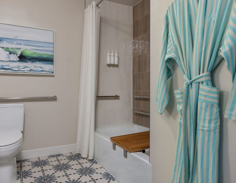 Accessible bathroom with a striped robe hanging on the wall, a toilet, walk-in shower with a bench, grab bars, and a curtain. A framed ocean wave picture and patterned tile floor are visible.