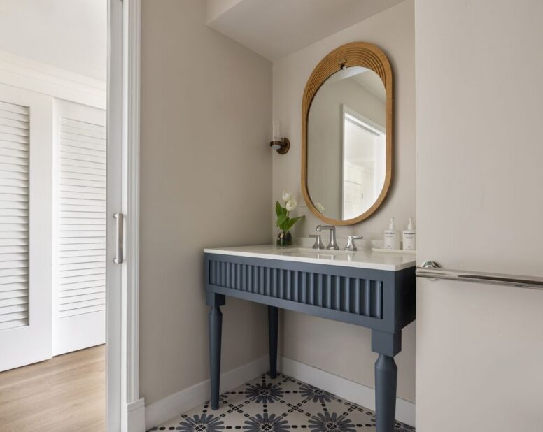 A modern bathroom with a blue vanity, oval mirror, single light fixture, white countertop, vase with white flowers, and geometric patterned floor tiles.