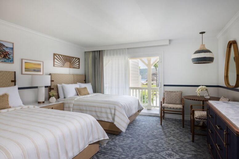 A bright hotel room with two double beds, striped bedding, a desk with two chairs, a round mirror, art on the walls, a pendant light, and a door leading to a sunny balcony.