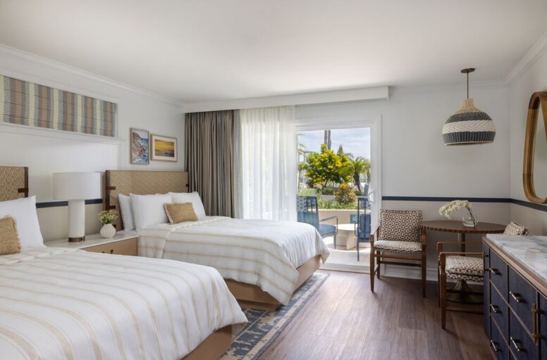 A bright hotel room with two beds, a seating area, and a dresser. Large window with sheer curtains opens to a balcony with outdoor seating and greenery. Decor is modern with neutral tones.