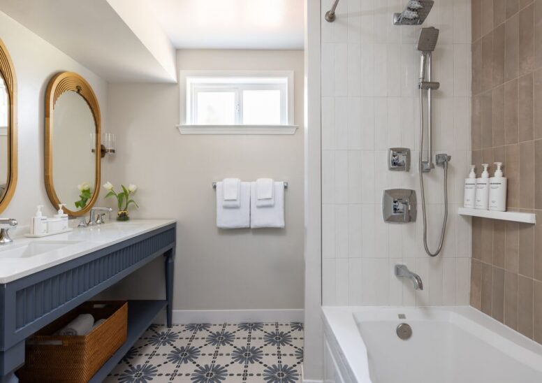 Modern bathroom with a double sink vanity, two mirrors, fresh flowers, towels on a rack, patterned tile floor, and a bathtub with both tan and white tiled walls and three bottles.