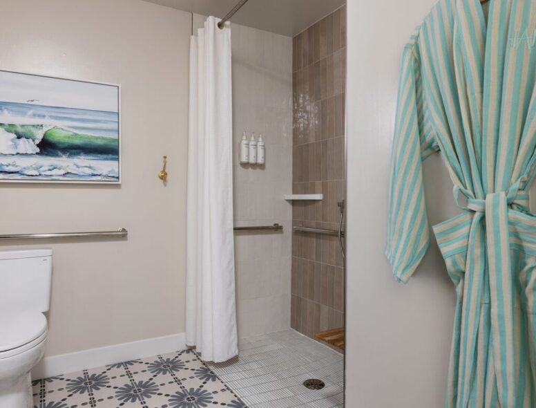 Bathroom with a walk-in shower, white curtain, grab bars, shampoo bottles, patterned floor tiles, surf art on the wall, and a green-striped robe hanging on the door.