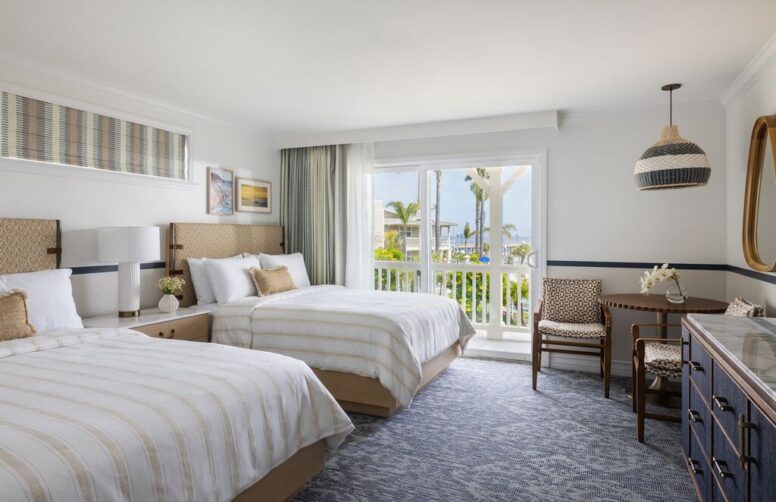 Hotel room with two double beds, a desk with two chairs, dresser, and balcony door. Light decor with blue accents, large window shows palm trees and ocean view outside.