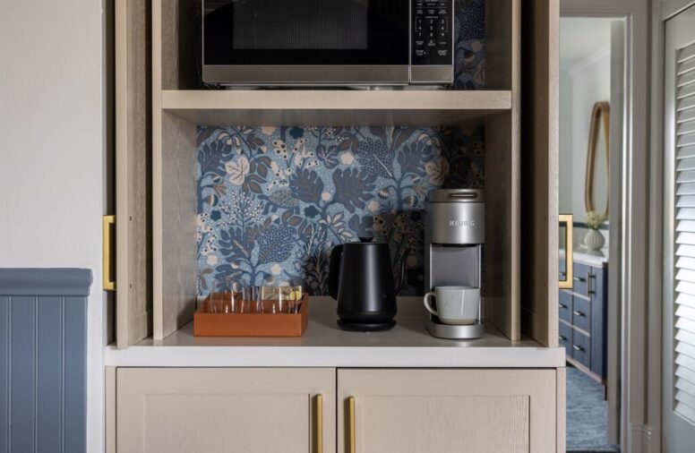 A small coffee station with a kettle, Keurig machine, mugs, and tea bags sits on a beige cabinet with gold handles, beneath a microwave and floral blue backsplash. A hallway and bathroom are visible in the background.