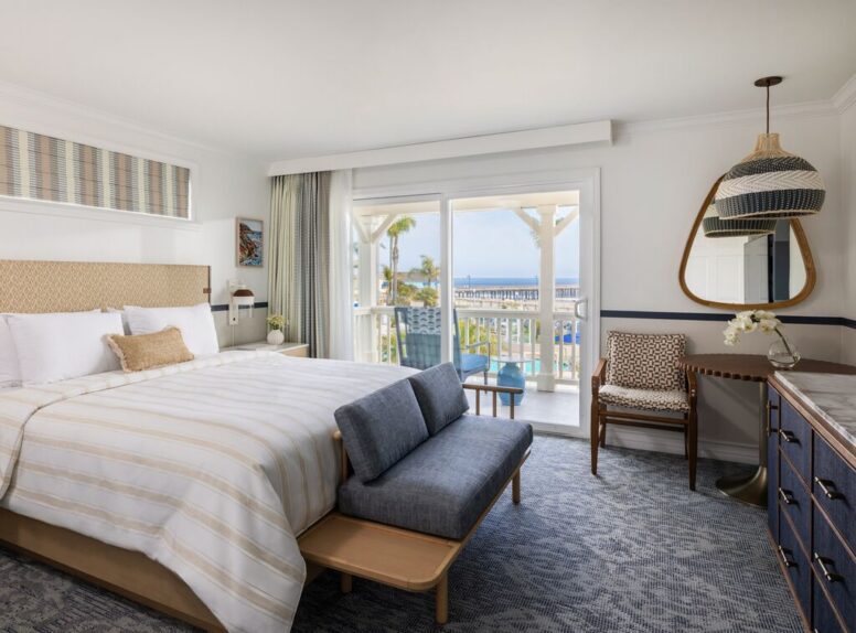 Bright hotel room with a king bed, blue-gray seating, dresser, mirror, and balcony; sliding doors reveal a view of palm trees and the ocean outside.