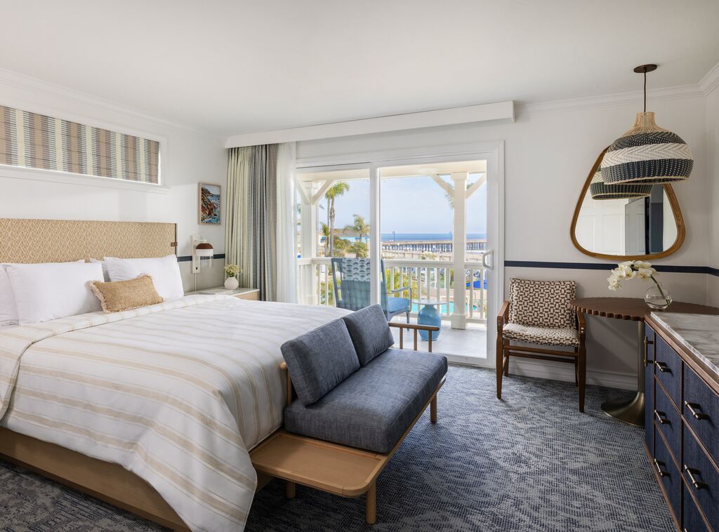 Bright hotel room with a king bed, blue-gray seating, dresser, mirror, and balcony; sliding doors reveal a view of palm trees and the ocean outside.