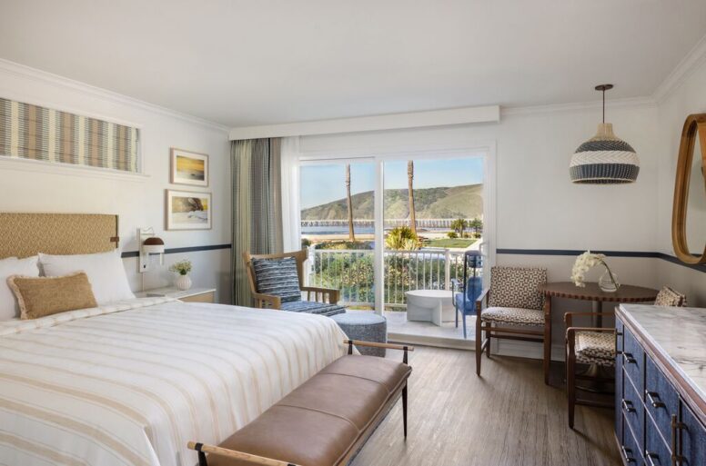 Bright hotel room with a king bed, seating area, and desk. Large window with a balcony overlooks green hills. Decor includes framed art, a round wall mirror, and light wood furniture.