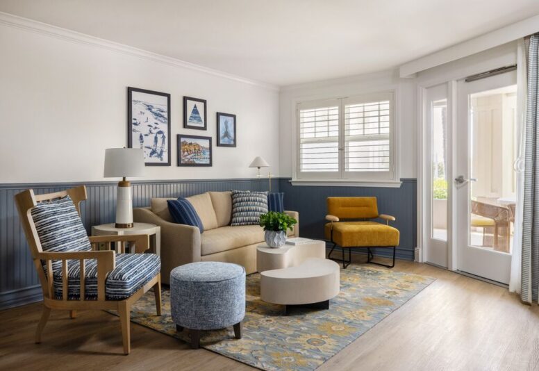 A cozy living room with a beige sofa, yellow armchair, wooden chair, round ottomans, rug, framed artwork on the wall, a side table with a lamp, and large windows with white shutters letting in natural light.