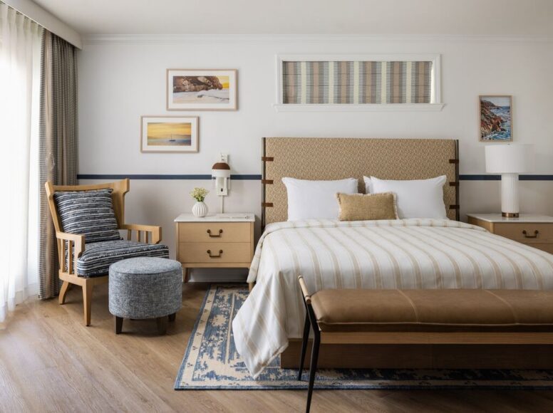 A cozy, modern bedroom with a large bed, beige headboard, wooden bench, armchair, ottoman, bedside tables, and framed art on the wall. Soft natural light filters through sheer curtains.