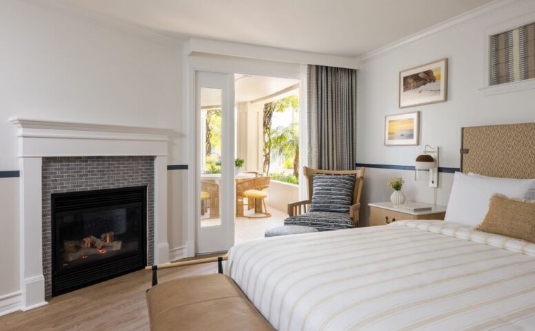 A cozy hotel room with a bed, fireplace, lounge chair, and sliding doors leading to a sunlit patio with a small table and chairs. Walls are white, with beach-themed art and greenery visible outside.