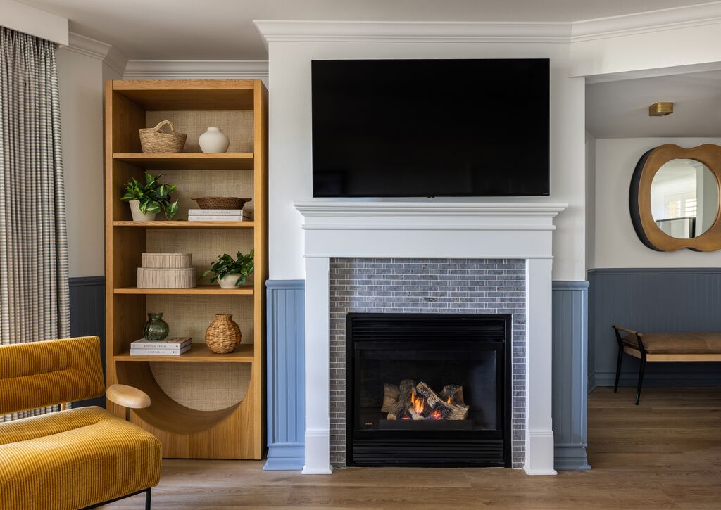 A modern living room with a fireplace, a large TV above it, a wooden bookshelf with decor items on the left, a mustard chair, wood floor, and a round mirror in the hallway on the right.