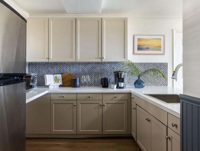 Modern kitchen with beige cabinets, stainless steel fridge, grey herringbone tile backsplash, appliances on the counter, a vase with a green plant, and a framed seascape picture on the wall.
