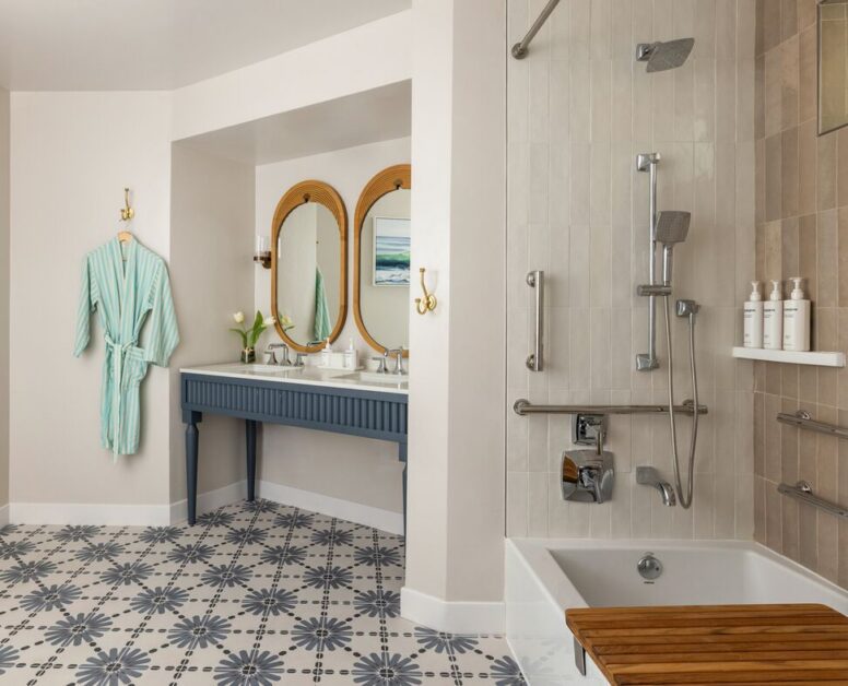 Modern bathroom with patterned tile floor, double sink with two mirrors, wall-mounted lights, a mint green robe, and a bathtub with a shower, grab bars, and wooden bench. White toiletries are arranged on a corner shelf.