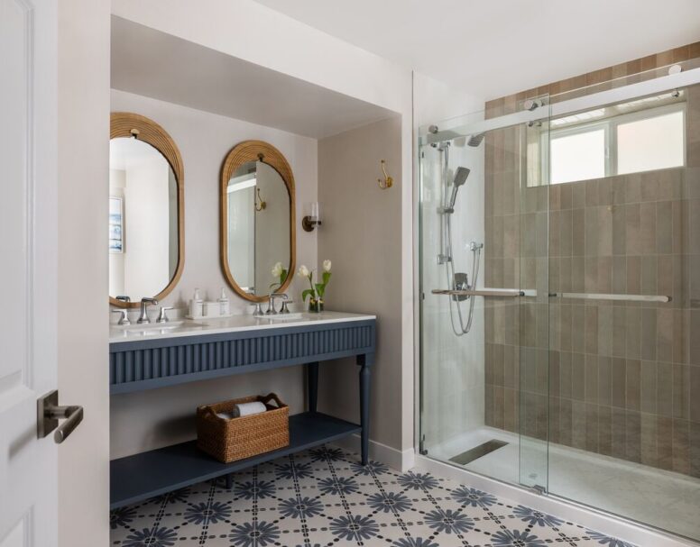 Modern bathroom with double sinks, oval mirrors, a blue vanity, a wicker basket, and a large glass-enclosed shower. The floor features patterned tiles, and a small vase with flowers sits on the counter.