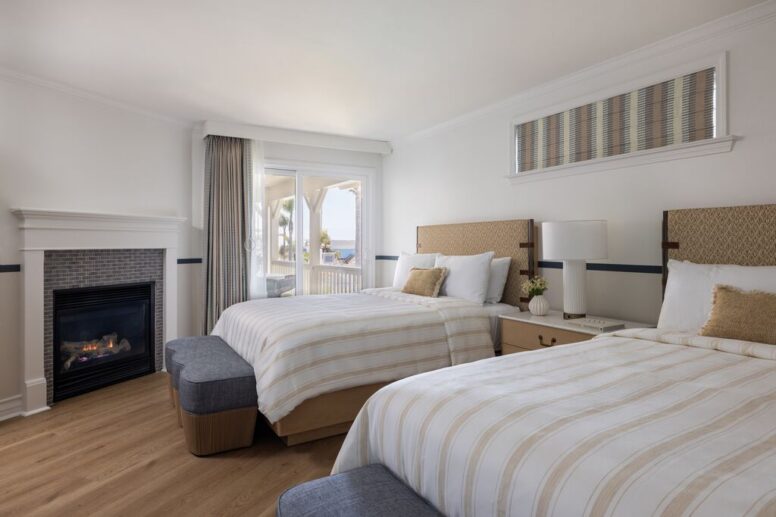 A bright hotel room with two double beds, a fireplace, nightstand with lamp, a large window with curtains, and a view of the ocean outside. The room has light wood floors and modern, neutral decor.