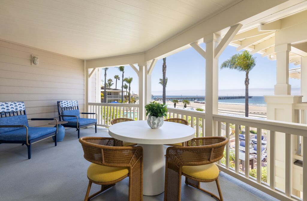 Covered patio with a round table, four chairs, two lounge chairs, and a potted plant, overlooking a beach and ocean with palm trees and a distant pier under a clear sky.