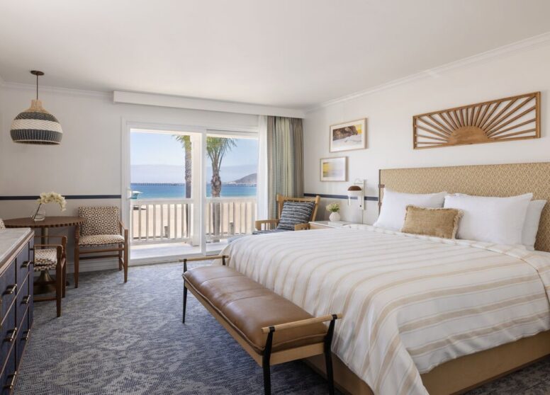 Bright hotel room with a large bed, bench, chairs, dresser, beach-themed decor, and a sliding glass door leading to a balcony with an ocean view and palm trees.
