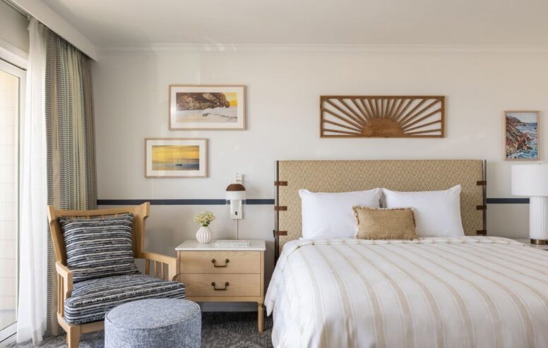 A neatly made bed with striped bedding, a cushioned headboard, a wooden chair with a striped pillow, a round ottoman, a nightstand with decor, and framed art on a white wall.