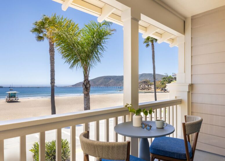 A cozy balcony overlooks a sandy beach with palm trees, blue ocean, distant pier, and hills. A small table with two chairs, coffee cups, and a plant invites relaxation under a bright, sunny sky.