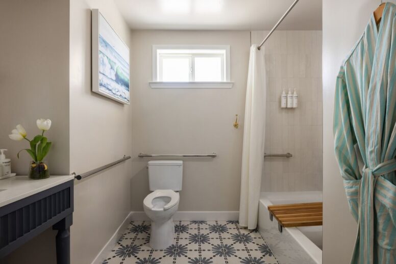 Accessible bathroom with patterned tile floor, toilet with grab bars, shower with curtain and seat, vanity with flowers, striped robe hanging, and a window above the toilet.