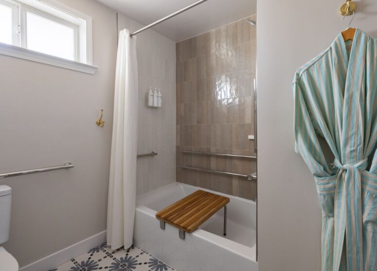 Accessible bathroom with a bathtub, shower curtain, grab bars, a wooden shower bench, patterned tile floor, and a striped robe hanging on the wall. There’s a window above the toilet.