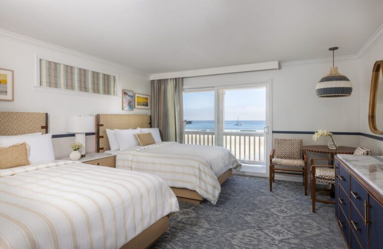 A bright hotel room with two double beds, a desk with chairs, wall art, a blue dresser, and a balcony overlooking the ocean with a sailboat in the distance.