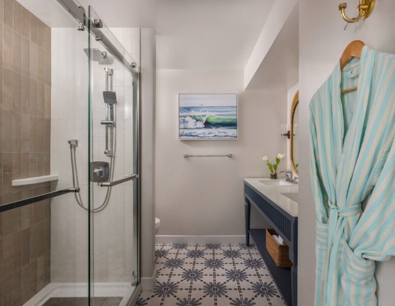Modern bathroom with a glass-enclosed shower, patterned tile floor, blue-striped robe on a hook, large vanity with a mirror and sink, and ocean artwork on the wall.