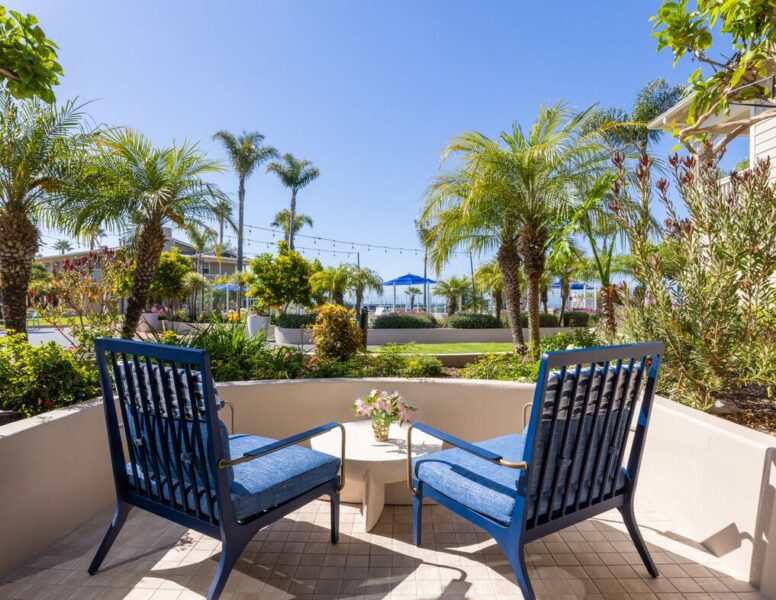 Two blue patio chairs and a small table with flowers sit on a tiled terrace, surrounded by palm trees and greenery, with blue umbrellas and string lights visible in the sunny background.