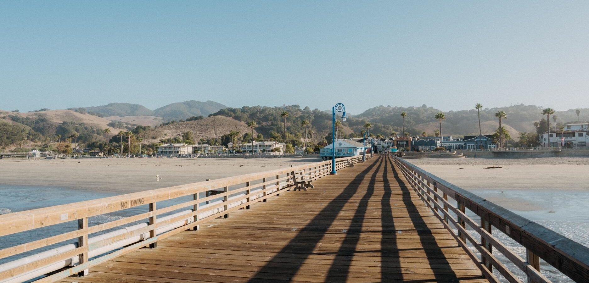 A wooden pier extends toward a beach with mountains, palm trees, and houses in the background under a clear blue sky. Shadows from the railings stretch across the pier in the sunlight.