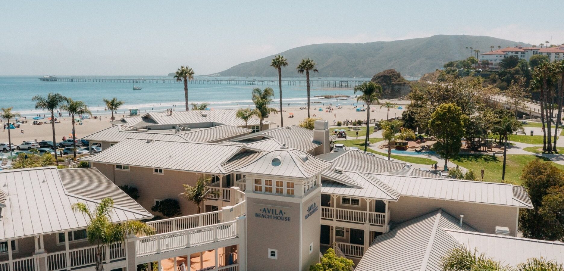 A coastal hotel labeled Avila Beach House overlooks a sandy beach with palm trees, people, parked cars, and a long pier; green hills rise in the background under a clear sky.