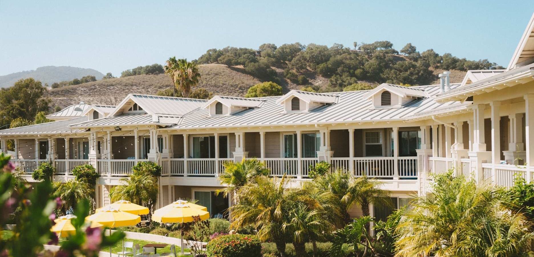 A two-story building with white railings and metal roofs, surrounded by palm trees and yellow patio umbrellas, set against hills and clear blue sky.