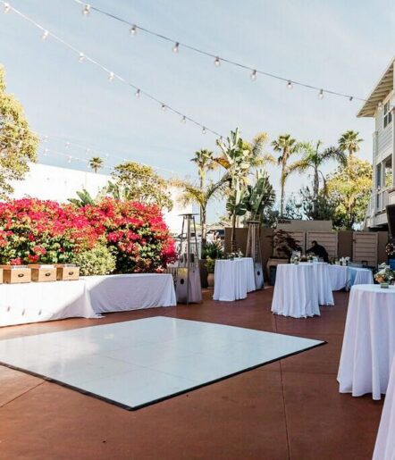 An outdoor event space with string lights, white tablecloth-covered tables, a white dance floor, vibrant flowers, palm trees, and clear skies.