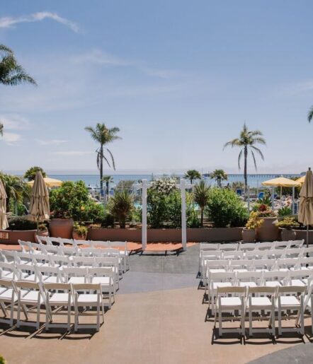 Rows of white chairs face a floral wedding arch on a patio with palm trees, overlooking the ocean and a pier under a clear blue sky.