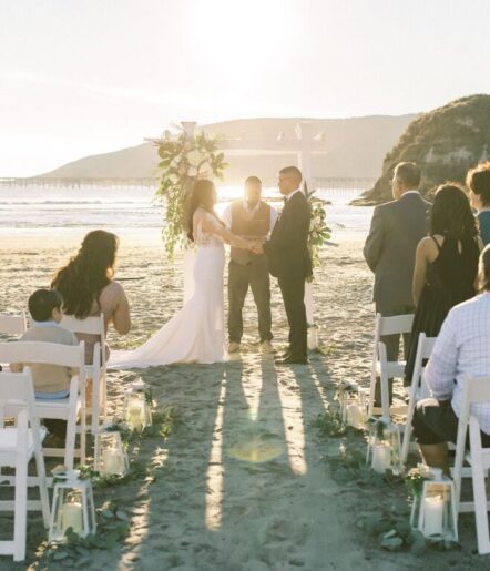 A bride and groom stand at an outdoor beach altar during sunset, holding hands as they exchange vows. Guests are seated on either side, and lanterns line the aisle. A pier and hills are visible in the background.