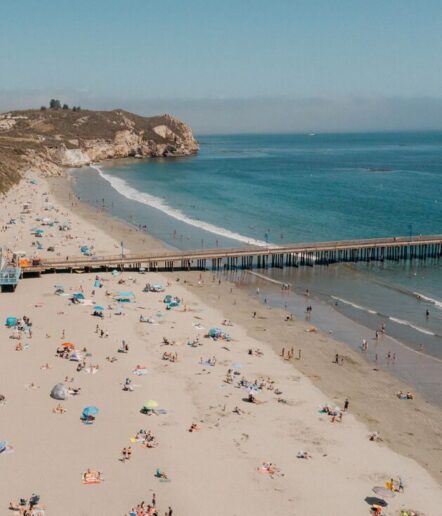 A crowded sandy beach with people sunbathing, swimming, and relaxing under umbrellas near a long pier. Hills and cliffs are visible in the background beside the blue ocean under a clear sky.