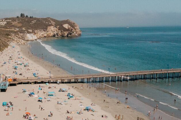 A sandy beach with scattered people and umbrellas, a long pier extending over calm blue water, and rocky cliffs in the background under a partly cloudy sky.