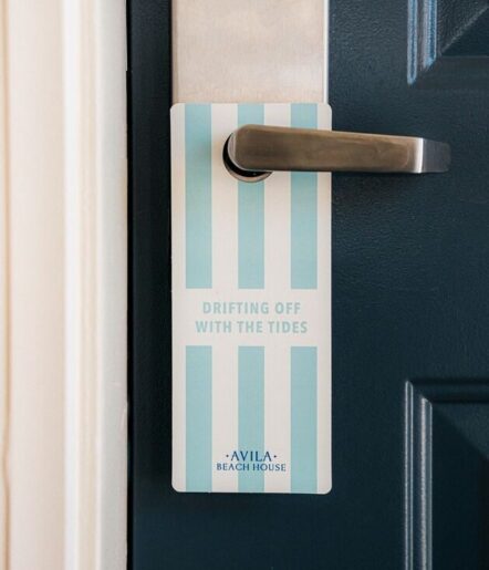 A door hanger with blue and white stripes hangs on a silver door handle. It reads “DRIFTING OFF WITH THE TIDES” and “AVILA BEACH HOUSE” at the bottom. The door is dark blue.